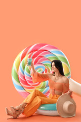 Wall Mural - Smiling young woman with cocktail, inflatable ring and mattress sitting on orange background