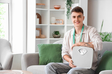 Poster - Young man with weight scales sitting on sofa at home