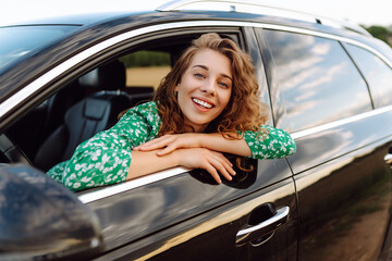 Wall Mural - A young traveler woman enjoying a sunny day by the roadside in a green dress, leaning out of a car window with a joyful expression. Summer trip. Lifestyle, travel, tourism, nature, active life