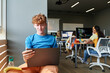 © Mediaphotos - Portrait of red haired young student relaxing by window in class and using laptop computer, copy space