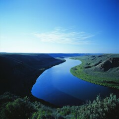 Poster - A scenic view of a winding river flowing through a valley, with green hills on either side and a clear blue sky above.