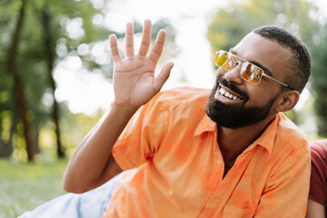 Smiling, happy African American man wearing sunglasses waving hand at someone in park looking away