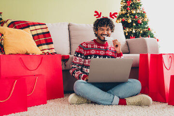 Wall Mural - Full body portrait of nice young man use laptop bite credit card eye wink wear xmas sweater holiday atmosphere flat indoors