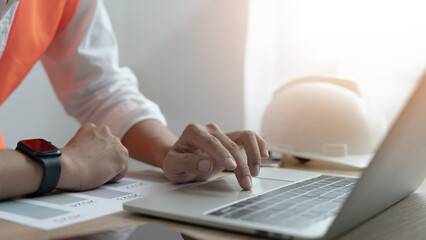 A person is typing on a laptop computer with a construction helmet on the desk. The scene suggests that the person is working on a construction project or related task