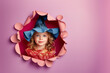© ZaStock - A cheerful girl wearing a vibrant witch hat smiles as she peers through a creatively torn pink wall, embracing the playful spirit of Halloween at a festive gathering