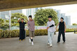 © DragonImages - Group of young men dancing energetically under urban overpass while wearing casual streetwear, surrounded by greenery and cityscape, enjoying outdoor activity together