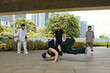 © DragonImages - Urban dancers showcasing breakdancing skills in park surrounded by tall buildings. Participants observing performance with trees and greenery in background