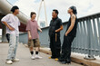 © DragonImages - Group of friends enjoying outdoor time near modern bridge structure engaging in conversation during their leisure time under sunny sky with cityscape background