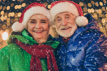 Canvas Print - Two retired pensioner grey white hair people affectionate couple woman man celebrate x-mas christmas jolly event walk outside under illumination wear santa claus headwear