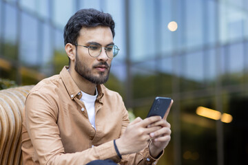 Young professional man sitting outside, engaging with smartphone. Wearing glasses and casual shirt, emphasizing modern digital lifestyle. Image conveys focus, communication, and internet connectivity