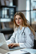 © ebhanu - A young female doctor therapist, dressed in a white medical uniform, is sitting at her desk in a medical office within a clinic. confident and focused as works on her laptop.