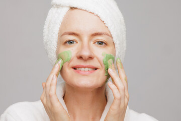 Joyful emotional woman applying green mask on face, facial skin care concept, cosmetology