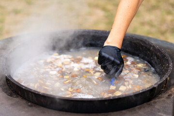 A hand wearing a black glove mixes ingredients in a large pot of stew, surrounded by a relaxing outdoor environment under bright sunlight.