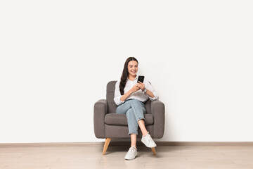 Wall Mural - Smiling woman with smartphone sitting on armchair against white wall