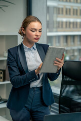 in modern office with a large window, a beautiful female worker stands in a blue suit with a tablet in her hand looking through the work plan for the month