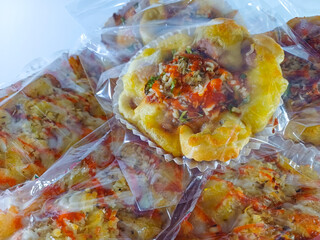 Stacks of cheesy bread topped with shredded cheese, chili sauce, and mayonnaise, neatly arranged in transparent plastic, set against a clean white background, highlighting their savory appeal