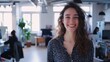 © VK Studio - A young woman with curly hair smiles brightly in a modern office space, filled with natural light and busy co-workers in the background.