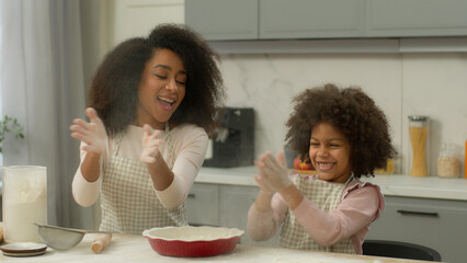 African American family make dough pizza pie pastry recipe cooking together mother woman teach little child kid girl daughter smile clapping dirty flour hands messy applauding palms cook at kitchen