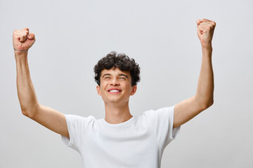 Happy young man celebrating success with raised arms and a joyful expression, wearing a white t shirt against a soft grey background, conveying positivity and achievement