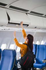 Wall Mural - Boarding Pass, Overhead Bin: A young woman stows her carry-on luggage in the overhead compartment of an airplane, ready for takeoff.