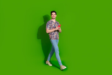 Poster - Full size photo of clever optimistic guy dressed print shirt jeans holding smartphone go empty space isolated on green color background