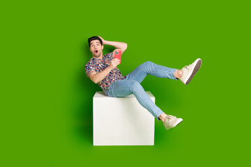 Canvas Print - Full size photo of astonished guy dressed print shirt sit on white cube holding smartphone arm on head isolated on green color background