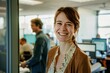 © Victor Bertrand - A woman with a bright smile stands in an open office space, exuding positivity and friendliness. Colleagues are seen working in the background.