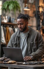 Black man in casual attire working on a laptop, with accessories on a table