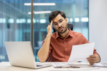 Wall Mural - Stressed businessman reviewing documents at office desk during workday, experiencing confusion and frustration