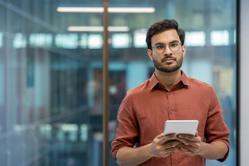 Confident Indian programmer standing in modern office holding tablet, showcasing professionalism and focus in tech industry.