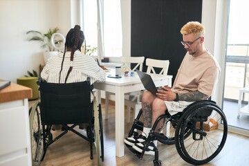 Two coworkers in wheelchairs working at desk with laptops and documents, collaborating on project in a modern office environment with large windows providing natural light