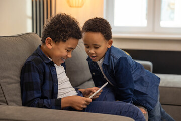 Cheerful boys friends playing with digital tablet while sitting on sofa