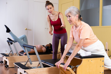 Wall Mural - Personal female trainer controlling movements of senior woman doing pilates on combo chair in fitness studio. Healthy active lifestyle