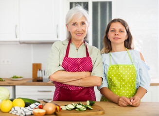 Wall Mural - Positive family, senior woman and her daughter in aprons, standing in kitchen in apartment. Preparing food at home.