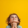 © Dai - A child with a curious expression, holding their chin and looking upwards, set against a bright yellow isolated background with clear space for adding text