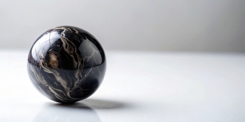 A single black marbled ball resting on a white surface, marble, ball, black, smooth, round, shiny, reflective, isolated