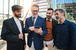 © Jose Calsina - A group of businessmen watching their investments on a mobile phone. Corporate males browsing the internet on a smartphone. Office workers displaying his phone to his business partner at a startup
