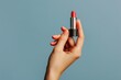 © kitipol - A womanâ€™s hand gracefully holding lipstick, cropped to highlight the elegance and femininity of the moment against a solid color background, blending style and poise
