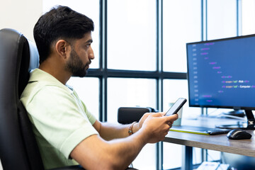 Using smartphone, Indian man sitting at desk with computer screen displaying code