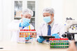 © NanSan - Two Asian scientists in lab coats sit at a table in a laboratory, examining samples through a microscope. The room is equipped with scientific tools, liquids, and chemistry materials.