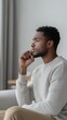 © Iswanto - A young man sits on a sofa, looking out of a window, with a thoughtful expression on his face. He is wearing a white sweater and khaki pants.