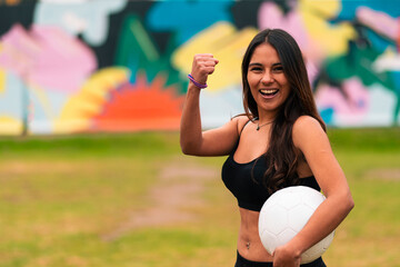 View of beautiful brunette female soccer player posing, holding and playing with soccer ball. On a green grass outdoors, with colorful background. sportswear. sports and wellness advertising