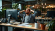 © altitudevisual - Man stretching with hands behind his head at desk in a modern office. Desk has a computer, telephone, notepads, and a plant. Open office environment in background.