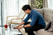 © Kittipong - Asian man wearing a long-sleeved shirt and long pants using a laptop on a sofa at home.