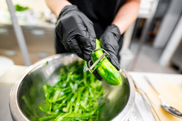 Close-up of hands in black gloves peeling cucumber into a stainless steel bowl filled with fresh green vegetable peels, in kitchen setting. Perfect for culinary, cooking, and food preparation themes.