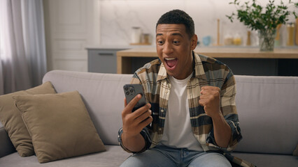 Wow African American man surprised wonder amazed win with phone mobile shocked sudden victory good winning news biracial male ethnic guy celebrating online success achieve offer excited on sofa couch