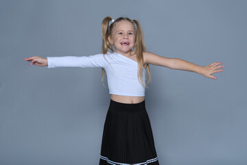 A cheerful young girl with blonde pigtails extends her arms wide, wearing a trendy white crop top and black pleated skirt. Her joyful expression conveys carefree childhood happiness.