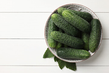 Fresh cucumbers in bowl and green leaves on white wooden table, top view. Space for text