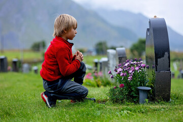 Sad little child, blond boy, standing in the rain on cemetery, sad person, mourning, rainy day