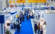 © Valentyna - Aerial view of crowded trade show floor with numerous blue-themed booths and attendees walking through wide aisles, showcasing a bustling business exhibition environment